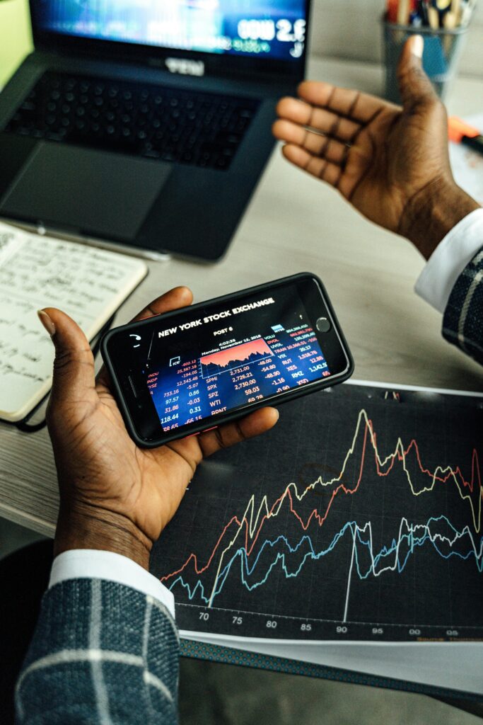 Close-up of hands holding a smartphone with stock market data, charts on paper and laptop in view.